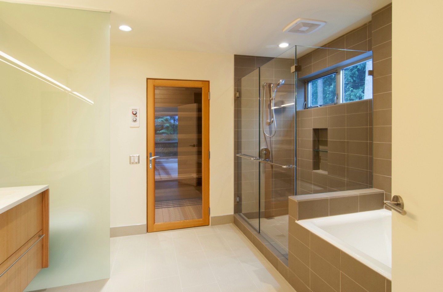 LaCantina(swing) A modern bathroom with a glass-enclosed shower, a bathtub, a wood door, a vanity with a sink, and a mirror. Walls are tiled in light brown, and the floor is light-colored.