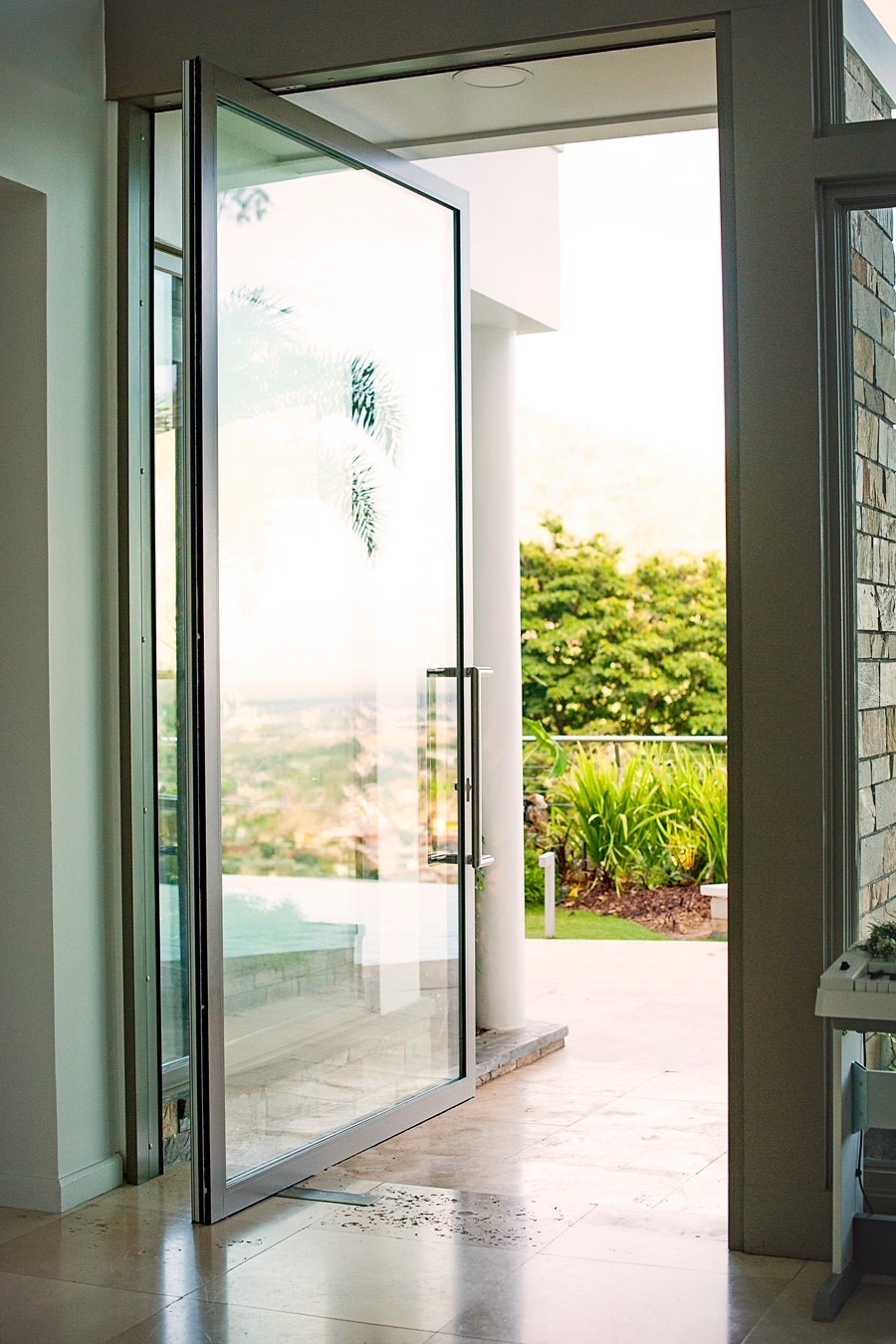 lacantinapivotdoor A glass door is open, leading to an outdoor area with lush greenery and partial view of a distant landscape. The interior has light-colored tile flooring.